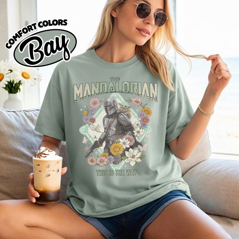 May include: A sage green short-sleeve t-shirt featuring a graphic of The Mandalorian and Grogu surrounded by colorful flowers. The text reads THE MANDALORIAN and THIS IS THE WAY. The shirt has a crew neckline and a relaxed fit design.