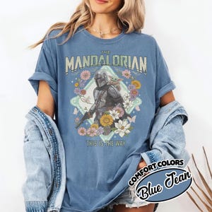 May include: A blue jean colored t-shirt featuring a graphic of The Mandalorian and Grogu surrounded by colorful flowers and butterflies. The text reads THE MANDALORIAN at the top and THIS IS THE WAY at the bottom in a vintage-style font.