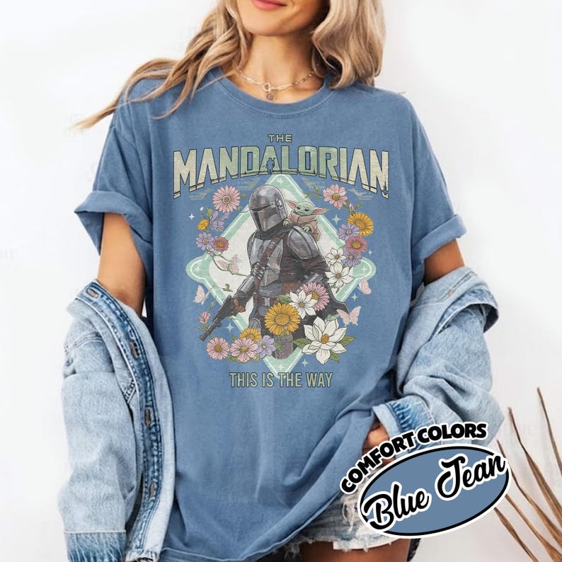 May include: A blue jean colored t-shirt featuring a graphic of The Mandalorian and Grogu surrounded by colorful flowers and butterflies. The text reads THE MANDALORIAN at the top and THIS IS THE WAY at the bottom in a vintage-style font.
