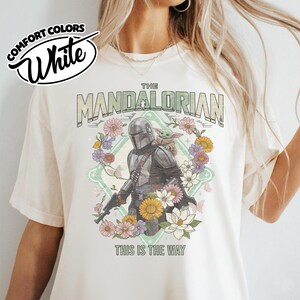 May include: A white t-shirt featuring a graphic of The Mandalorian and Grogu surrounded by a floral wreath. The text reads THE MANDALORIAN and THIS IS THE WAY. A circular logo in the top left corner displays the text COMFORT COLORS White.