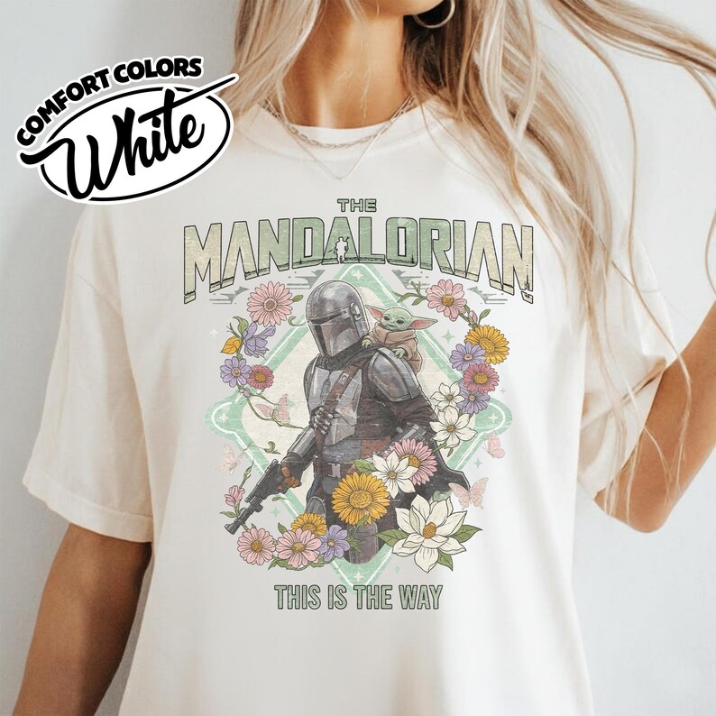 May include: A white t-shirt featuring a graphic of The Mandalorian and Grogu surrounded by a floral wreath. The text reads THE MANDALORIAN and THIS IS THE WAY. A circular logo in the top left corner displays the text COMFORT COLORS White.