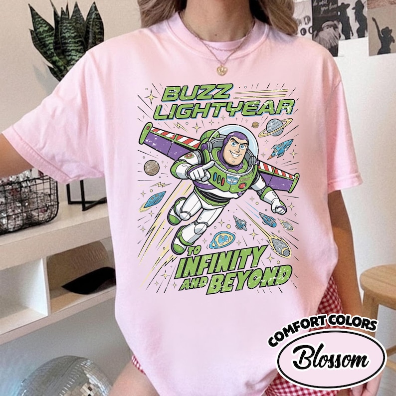 May include: A light pink t-shirt featuring a graphic of Buzz Lightyear flying with wings extended. The text reads BUZZ LIGHTYEAR TO INFINITY AND BEYOND. The design includes planets, stars, and small spacecraft in shades of green, purple, and blue.