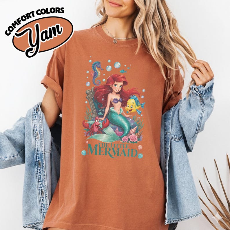 May include: Rust-colored t-shirt featuring a colorful graphic of Ariel from The Little Mermaid, with Flounder, Sebastian, and a seahorse. The text "THE LITTLE MERMAID" is below the graphic. The shirt has the text "COMFORT COLORS" and "Yam" in a brown oval.