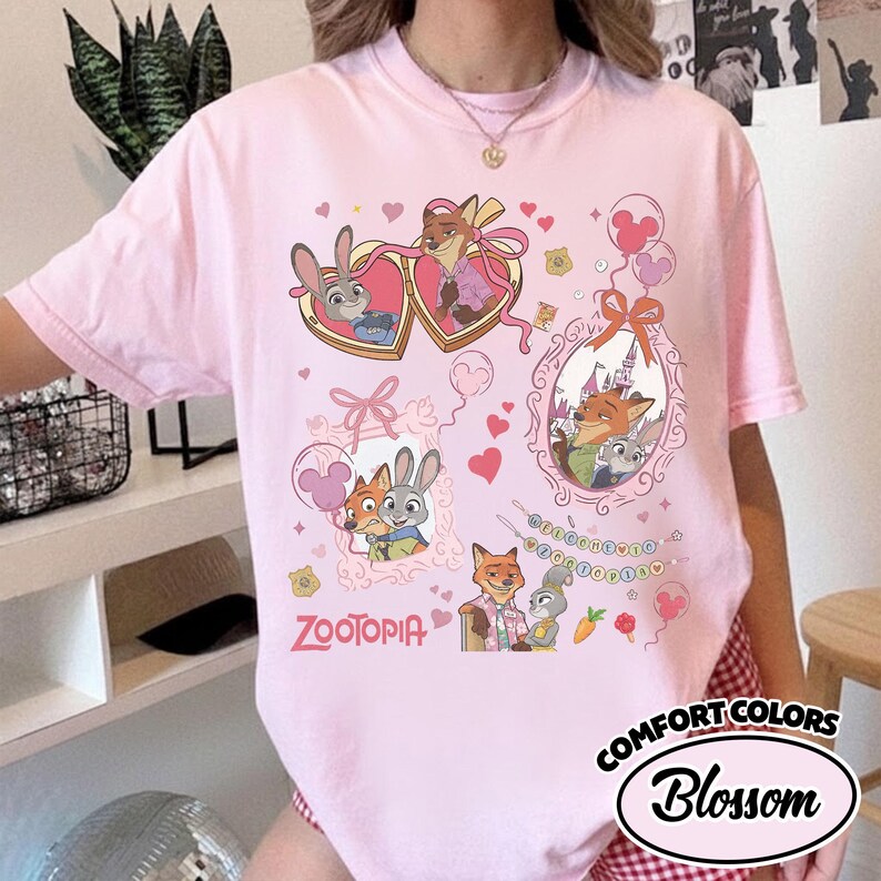 May include: A light pink t-shirt featuring a graphic print of characters from Zootopia, including Judy Hopps and Nick Wilde. The design includes heart-shaped frames, balloons, a string of beads spelling Zootopia, and the Zootopia logo in pink.