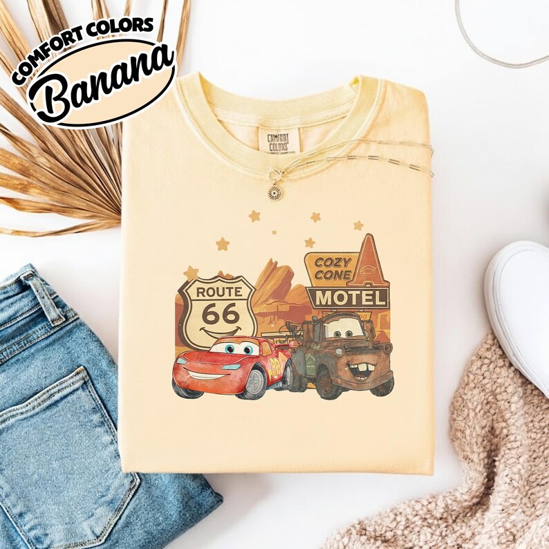 May include: A light yellow sweatshirt with a graphic featuring characters from the movie Cars, including Lightning McQueen and Mater, with a Route 66 sign and the words "Cozy Cone Motel". The text "Comfort Colors Banana" is printed on the top left.