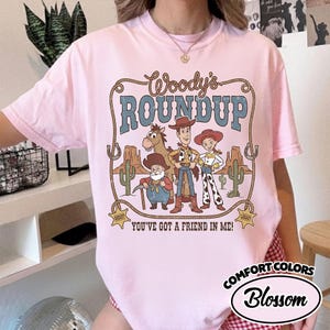 May include: A light pink t-shirt featuring a graphic of Woody's Roundup characters. The design includes the text "Woody's Roundup" in a brown, rope-style font, and the phrase "You've Got a Friend in Me!" in a brown font. The shirt also has the words "Comfort Colors" and "Blossom".