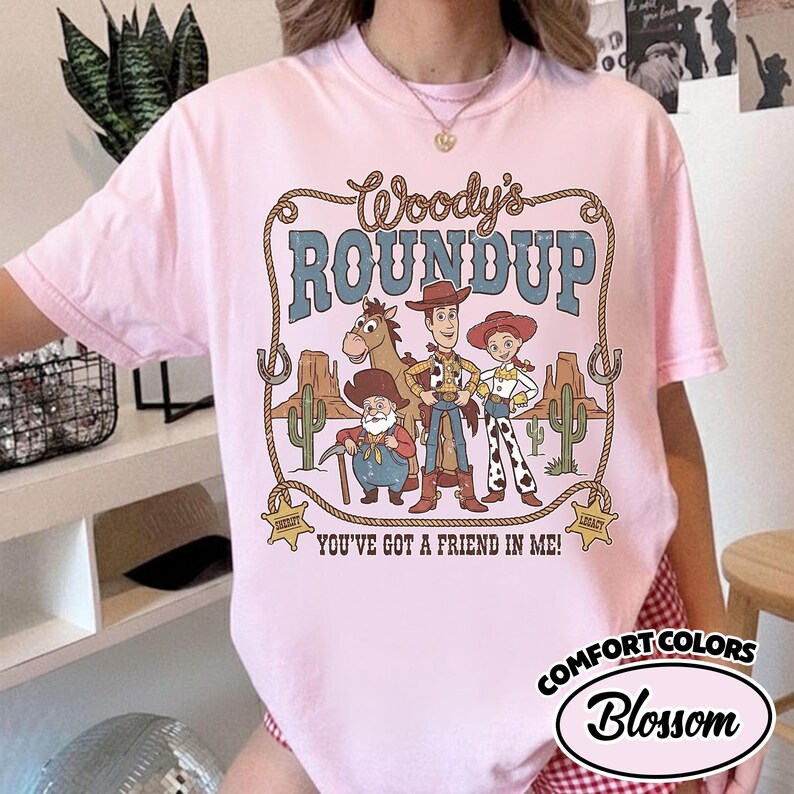 May include: A light pink t-shirt featuring a graphic of Woody's Roundup characters. The design includes the text "Woody's Roundup" in a brown, rope-style font, and the phrase "You've Got a Friend in Me!" in a brown font. The shirt also has the words "Comfort Colors" and "Blossom".