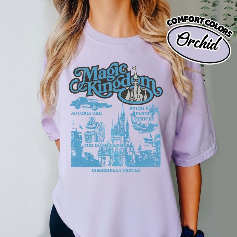 May include: A light purple t-shirt with a graphic featuring the words "Magic Kingdom" in a retro font. The design includes images of Autopia Car, Peter Pan's Flight Vehicle, Tiki Room Drummer, and Cinderella Castle. The text "Comfort Colors" and "Orchid" are also visible.