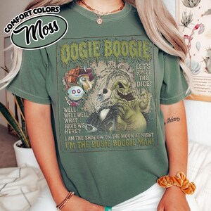 May include: Moss green t-shirt featuring a graphic from the movie "The Nightmare Before Christmas." The design includes the text "Oogie Boogie" and other phrases. The shirt has short sleeves and a crew neck.