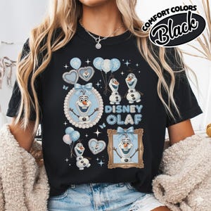 May include: Black t-shirt featuring a Disney Olaf design. The graphic includes Olaf characters, balloons, and the text "Disney Olaf" in blue. The shirt has a crew neck and short sleeves. The words "Comfort Colors Black" are printed on the upper right.