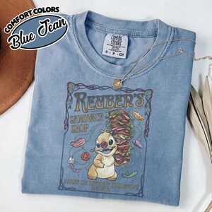 K&ouml;nnte beinhalten: Ein blaues Comfort Colors T-Shirt mit einer Grafik f&uuml;r "Reuben's Sandwich Shop", das eine Zeichentrickfigur und ein gro&szlig;es Sandwich zeigt. Das Shirt hat einen Rundhalsausschnitt und kurze &Auml;rmel. Eine goldene Halskette ist &uuml;ber dem Shirt drapiert.