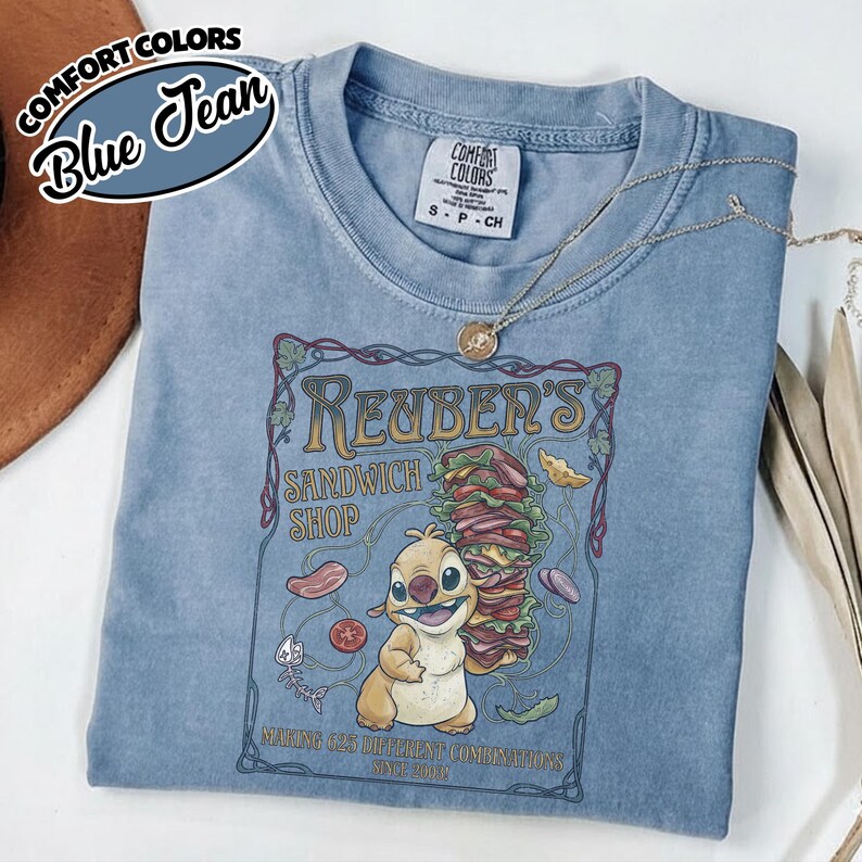K&ouml;nnte beinhalten: Ein blaues Comfort Colors T-Shirt mit einer Grafik f&uuml;r "Reuben's Sandwich Shop", das eine Zeichentrickfigur und ein gro&szlig;es Sandwich zeigt. Das Shirt hat einen Rundhalsausschnitt und kurze &Auml;rmel. Eine goldene Halskette ist &uuml;ber dem Shirt drapiert.