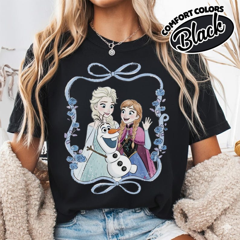 May include: Black t-shirt featuring a cartoon image of Elsa, Anna, and Olaf from the movie Frozen, framed by a blue ribbon and flowers. The words "COMFORT COLORS" and "Black" are printed on the upper right.