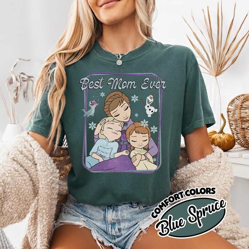 May include: A teal Comfort Colors t-shirt with a graphic that says "Best Mom Ever." The graphic features a mother and two children, with snowflakes and characters from the movie Frozen. The shirt is labeled "Blue Spruce."