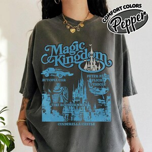 May include: A gray t-shirt with a blue graphic featuring the words "Magic Kingdom" and illustrations of Cinderella Castle, Autopia Car, Peter Pan's Flight Vehicle, and Tiki Room. The shirt also has the text "COMFORT COLORS" and "Pepper".