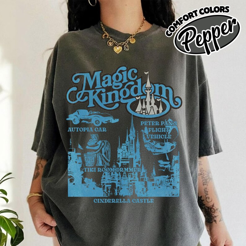 May include: A gray t-shirt with a blue graphic featuring the words "Magic Kingdom" and illustrations of Cinderella Castle, Autopia Car, Peter Pan's Flight Vehicle, and Tiki Room. The shirt also has the text "COMFORT COLORS" and "Pepper".