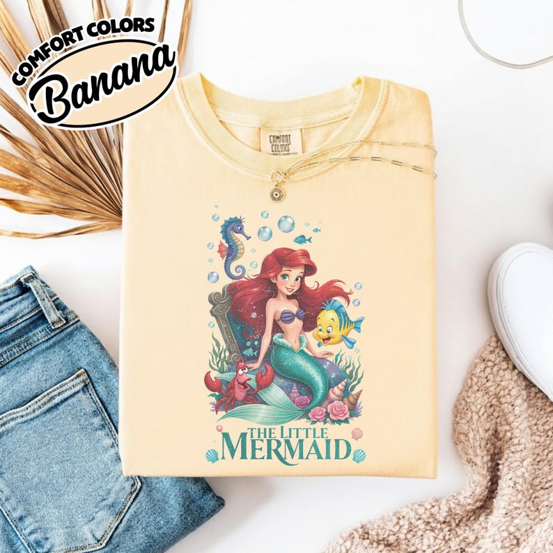 May include: A light yellow t-shirt featuring a graphic of Ariel from The Little Mermaid, with the text "The Little Mermaid". The shirt also has the text "Comfort Colors" and "Banana" in a circular design.
