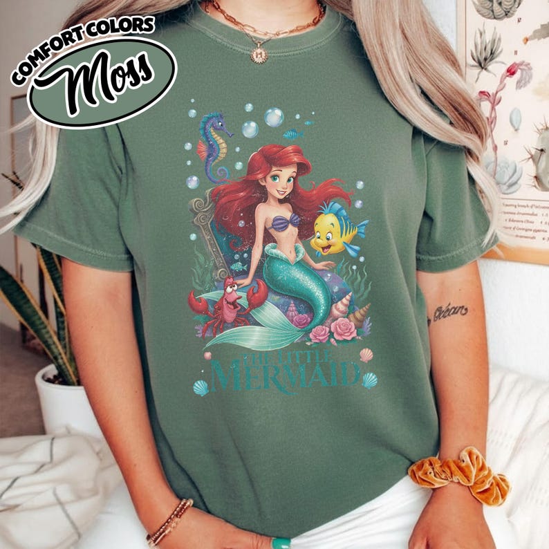 May include: Moss green t-shirt featuring a colorful graphic of Ariel from The Little Mermaid. The design includes Ariel, Flounder, Sebastian, and other sea creatures. The words "THE LITTLE MERMAID" are printed below the graphic. The shirt also has the words "COMFORT COLORS" and "Moss" printed on it.
