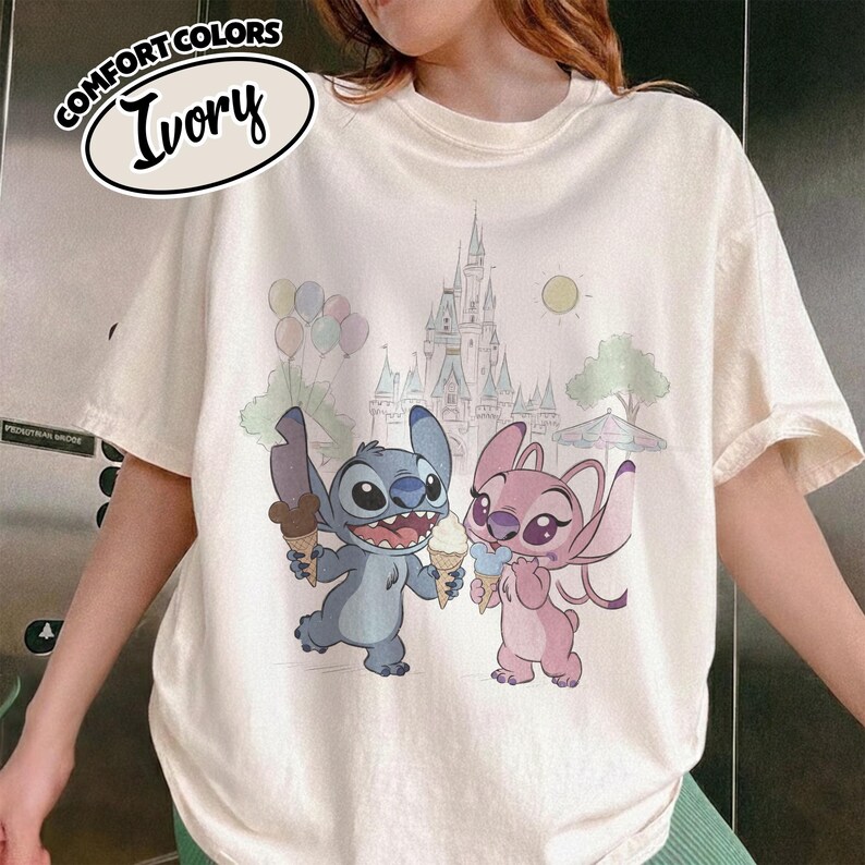 Puede incluir: Camiseta de color marfil con una ilustraci&oacute;n de dibujos animados de Stitch y Angel sosteniendo conos de helado frente a un castillo y globos. La camiseta tiene las palabras "COMFORT COLORS" e "Ivory" en un dise&ntilde;o circular.