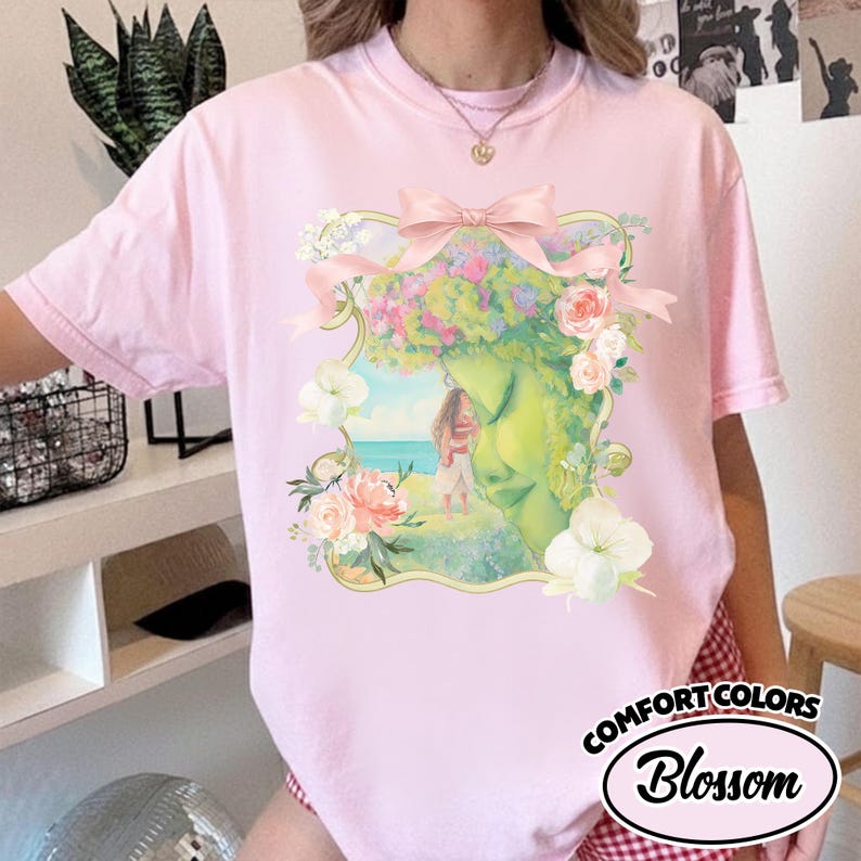 K&ouml;nnte beinhalten: Hellrosa T-Shirt mit einem floralen und Gesichts-Design, verziert mit einer rosa Schleife. Die Grafik zeigt eine Frau auf einem grasbewachsenen H&uuml;gel mit dem Meer im Hintergrund. Der Text "Comfort Colors Blossom" ist unten rechts aufgedruckt.