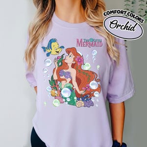 K&ouml;nnte beinhalten: Ein hellviolettes Kurzarm-T-Shirt mit einer Grafik von Arielle, die Meerjungfrau, und Fabius, umgeben von Blasen, Korallen und Muscheln. Der Text The Little Mermaid ist in einer rosa Schrift &uuml;ber der Figur aufgedruckt.