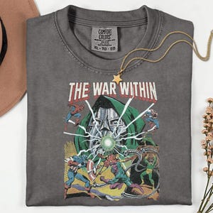 May include: A gray Comfort Colors t-shirt featuring a comic book graphic with the text "THE WAR WITHIN." The design includes illustrations of superheroes and a villain. A gold star pendant necklace rests on the shirt.