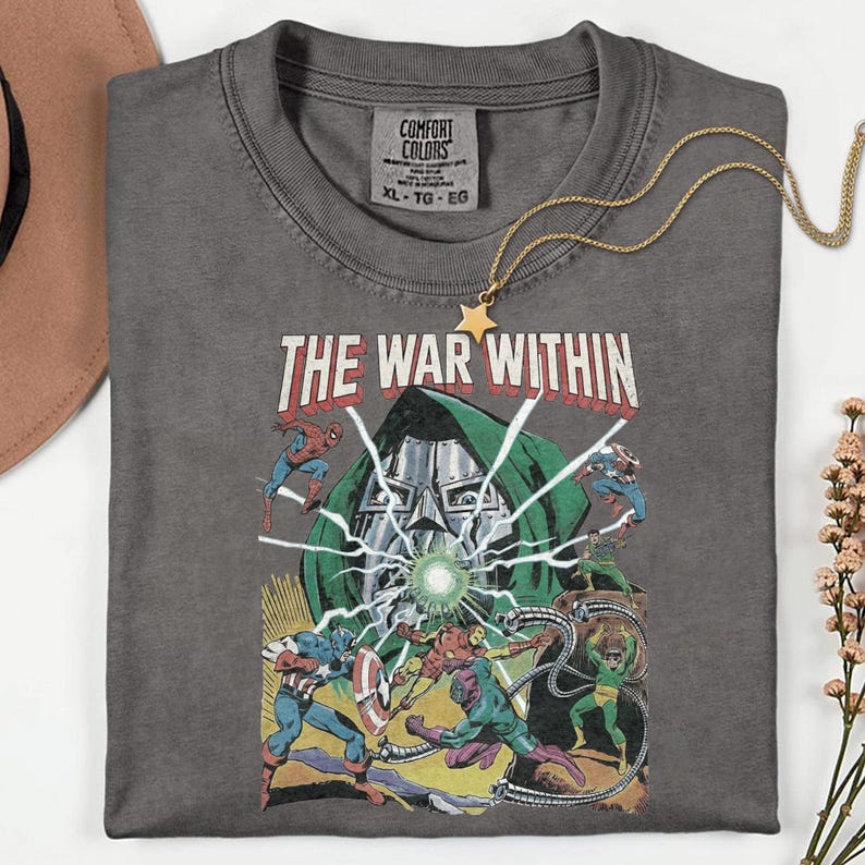 May include: A gray Comfort Colors t-shirt featuring a comic book graphic with the text "THE WAR WITHIN." The design includes illustrations of superheroes and a villain. A gold star pendant necklace rests on the shirt.