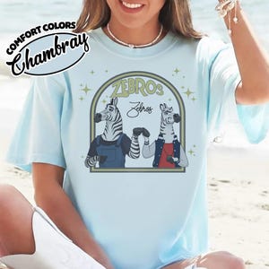 May include: Pale blue t-shirt featuring the word "ZEBROS" and a graphic of two zebras. One zebra is in overalls, the other in a jacket. The shirt also has the words "Comfort Colors" and "Chambray" in a circular design.
