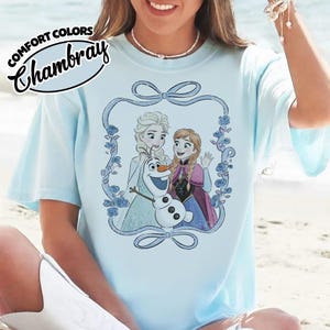 May include: Light blue t-shirt featuring a graphic of Elsa, Anna, and Olaf from the movie Frozen, framed by a blue floral design. The shirt has the words "COMFORT COLORS Chambray" printed on the top left.