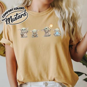 以下が含まれることがあります： 人気のSFシリーズのキャラクターの4つのイラストが描かれたマスタード色のTシャツ。キャラクターは、バッグ、テディベア、星、宝石を持っているさまざまなポーズで描かれています。「COMFORT COLORS」と「Mustard」の文字が左上にプリントされています。