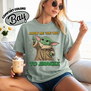 May include: Sage green t-shirt with a graphic of a green alien character pointing upwards, with the text "SHOW ME THE WAY TO SNACKS". The shirt also features the Comfort Colors Bay logo.