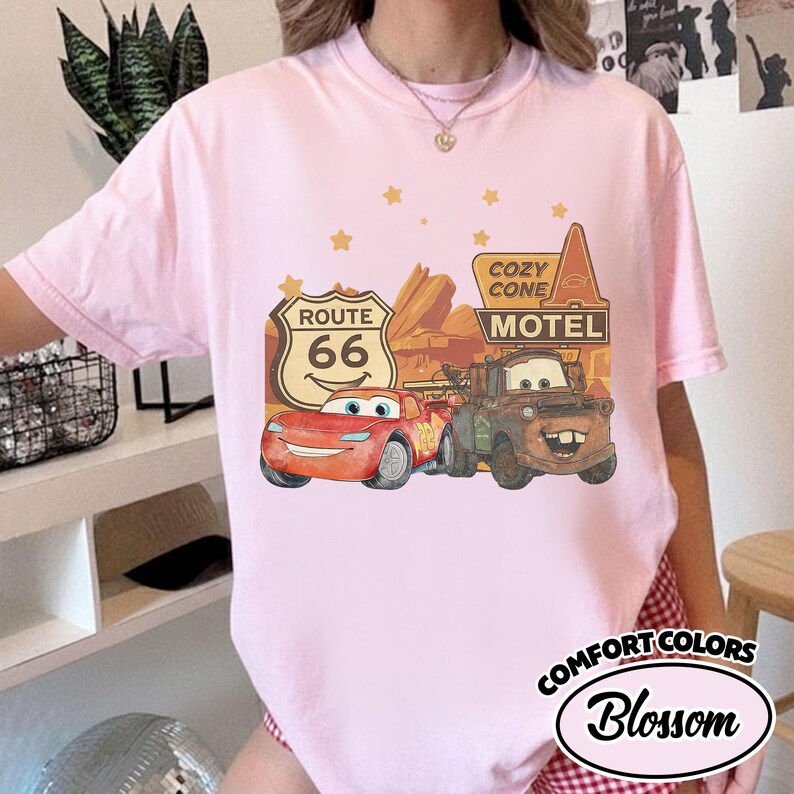 May include: A light pink t-shirt featuring a graphic of the Cars movie characters Lightning McQueen and Mater, along with Route 66 and Cozy Cone Motel signs. The shirt also has the words "Comfort Colors" and "Blossom" printed on it.