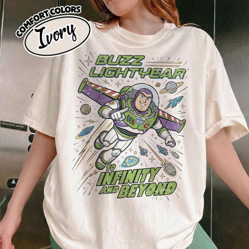 May include: An ivory t-shirt featuring a graphic of Buzz Lightyear in a space suit with wings. The text reads BUZZ LIGHTYEAR TO INFINITY AND BEYOND. The design includes planets, stars, and small spacecraft in shades of green, purple, and blue.