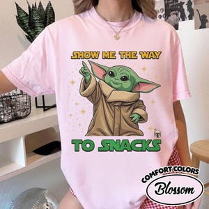 May include: A light pink Comfort Colors t-shirt featuring a graphic of a green alien character pointing upwards. The text "SHOW ME THE WAY TO SNACKS" is printed above and below the character in yellow and green.