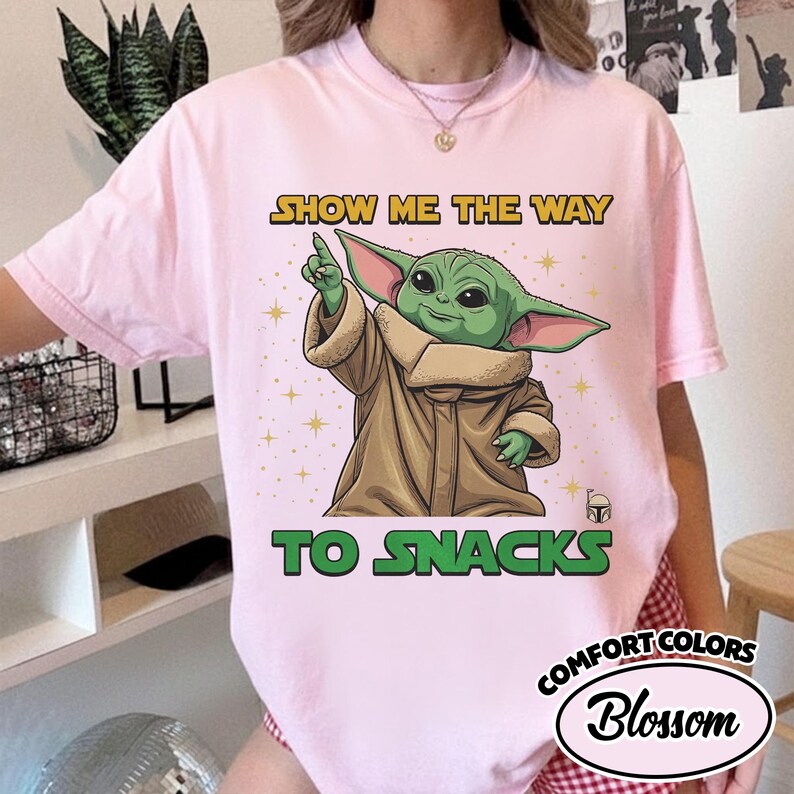 May include: A light pink Comfort Colors t-shirt featuring a graphic of a green alien character pointing upwards. The text "SHOW ME THE WAY TO SNACKS" is printed above and below the character in yellow and green.