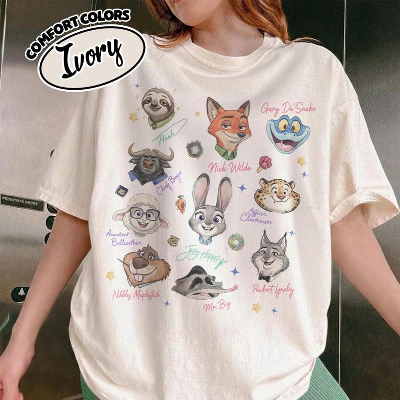 K&ouml;nnte beinhalten: Elfenbeinfarbenes Comfort Colors T-Shirt mit Illustrationen von Zeichentrickfiguren. Das Design umfasst Charaktere wie Nick Wilde, Judy Hopps und Flash. Der Text "COMFORT COLORS" und "Ivory" ist ebenfalls sichtbar.