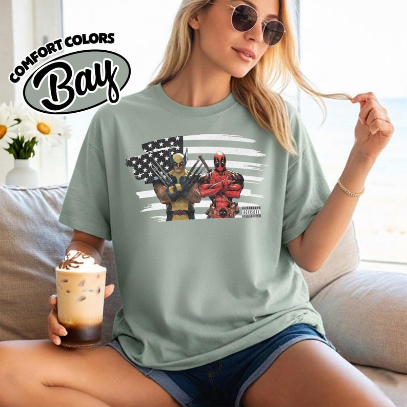 May include: A sage green short-sleeve t-shirt featuring a graphic print of Wolverine and Deadpool standing in front of a stylized American flag. The shirt includes a small black rectangular label with the text Parental Advisory.