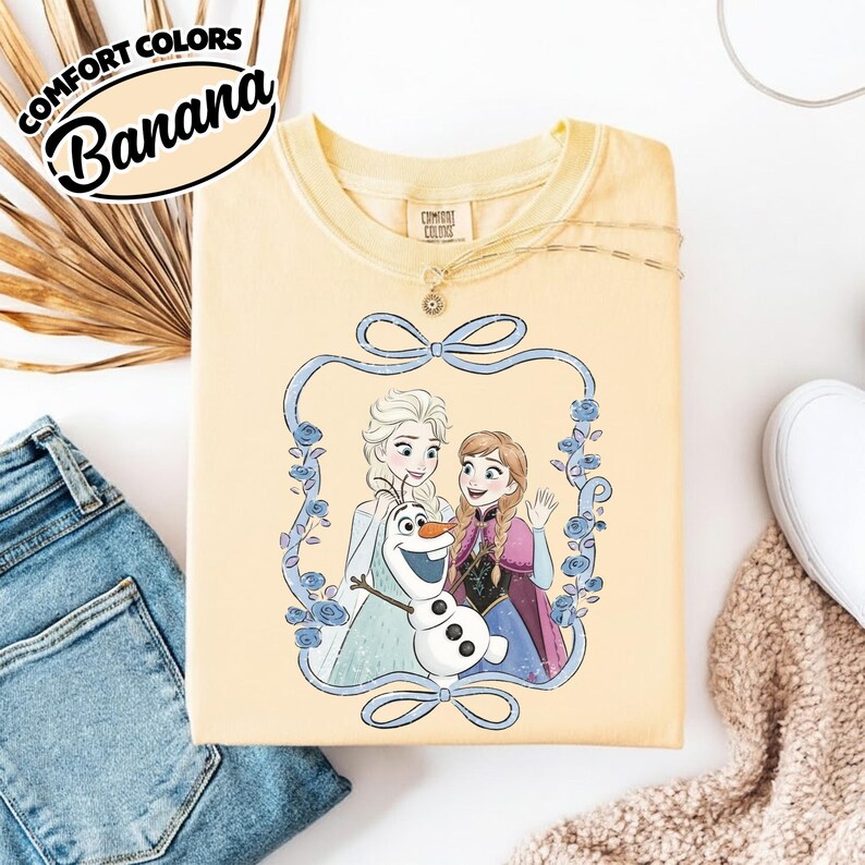 May include: A light yellow Comfort Colors sweatshirt featuring a graphic of Elsa, Anna, and Olaf from Frozen, framed by a blue ribbon and floral design. The text "Comfort Colors Banana" is printed at the top left.