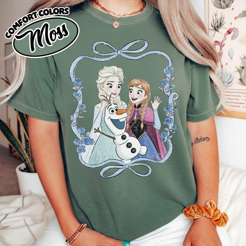 May include: Moss green t-shirt featuring a graphic of Elsa, Anna, and Olaf from Frozen, framed by a blue ribbon and roses. The words "COMFORT COLORS" and "Moss" are in a circular design at the top left.