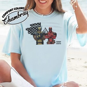 May include: A light blue t-shirt featuring a graphic print of Wolverine and Deadpool standing in front of a stylized American flag. The design includes the text Comfort Colors Chambray and a small Parental Advisory label in the bottom right corner.
