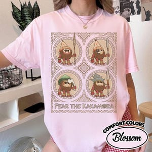 May include: Pale pink Comfort Colors t-shirt with a graphic of four cartoon figures in circles, each holding a spear. The text "FEAR THE KAKAMORA" is below the graphic. The shirt also has the text "Comfort Colors Blossom".
