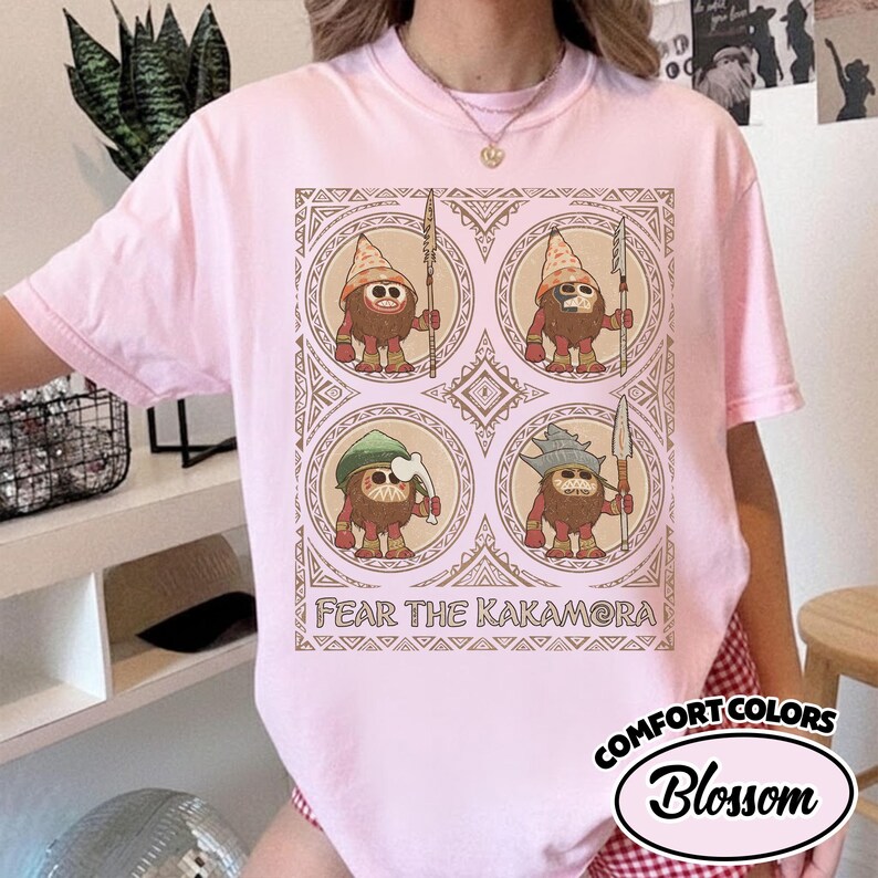 May include: Pale pink Comfort Colors t-shirt with a graphic of four cartoon figures in circles, each holding a spear. The text "FEAR THE KAKAMORA" is below the graphic. The shirt also has the text "Comfort Colors Blossom".
