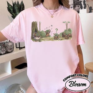 May include: Light pink t-shirt featuring a cartoon illustration of a character from a popular science fiction franchise. The graphic shows several of the character in a forest setting. The shirt has the text "COMFORT COLORS" and "Blossom" in a circular design.