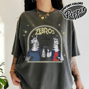 May include: Dark grey t-shirt with a cartoon illustration of two zebras bumping fists. The design includes the word "ZEBROS" in a yellow arch, with the word "Zebros" in script below. The shirt also has the text "COMFORT COLORS Pepper".