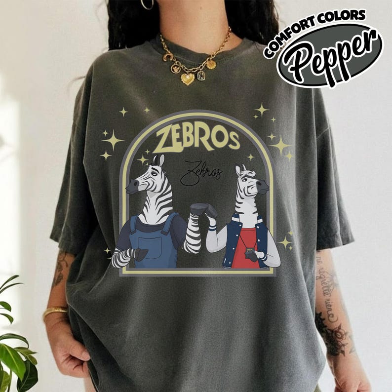 May include: Dark grey t-shirt with a cartoon illustration of two zebras bumping fists. The design includes the word "ZEBROS" in a yellow arch, with the word "Zebros" in script below. The shirt also has the text "COMFORT COLORS Pepper".