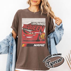 May include: A dark brown short-sleeve t-shirt featuring a graphic of the red race car Lightning McQueen. The design includes the text "LIGHTNING MCQUEEN" in bold red letters and "NAME" in white, with a yellow, orange, and red stripe accent.