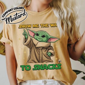 May include: Mustard-colored t-shirt featuring a cartoon illustration of a green alien character pointing upwards. The text "SHOW ME THE WA TO SNACKS" is printed in yellow and green. The shirt has a relaxed fit.