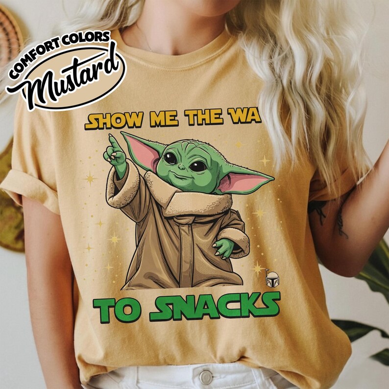 May include: Mustard-colored t-shirt featuring a cartoon illustration of a green alien character pointing upwards. The text "SHOW ME THE WA TO SNACKS" is printed in yellow and green. The shirt has a relaxed fit.