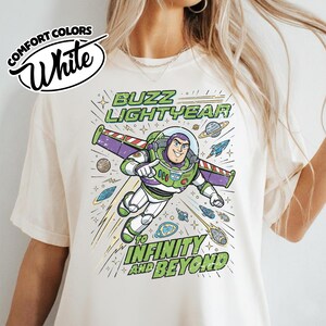 May include: A cream-colored t-shirt featuring a graphic of Buzz Lightyear in a space suit with wings extended. The text reads BUZZ LIGHTYEAR TO INFINITY AND BEYOND. The design includes planets, stars, and small spaceships in blue, green, and purple.