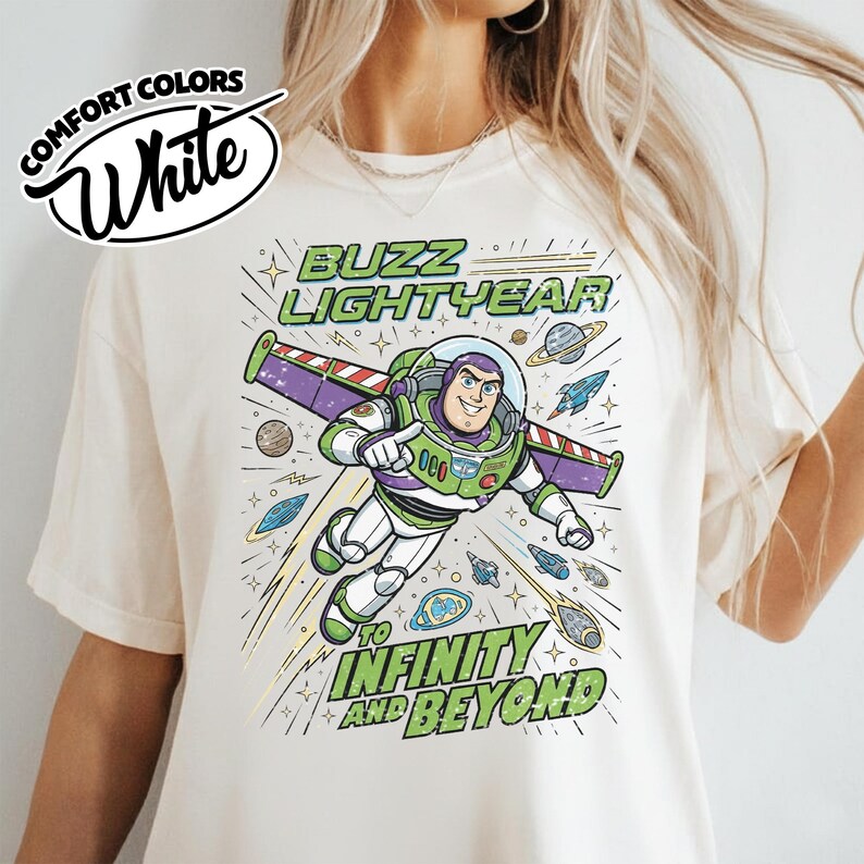 May include: A cream-colored t-shirt featuring a graphic of Buzz Lightyear in a space suit with wings extended. The text reads BUZZ LIGHTYEAR TO INFINITY AND BEYOND. The design includes planets, stars, and small spaceships in blue, green, and purple.